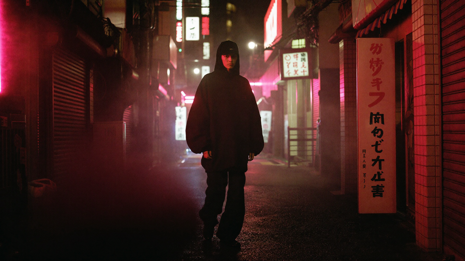 Model in an oversized black hoodie in a neon-lit Tokyo back-alley at midnight